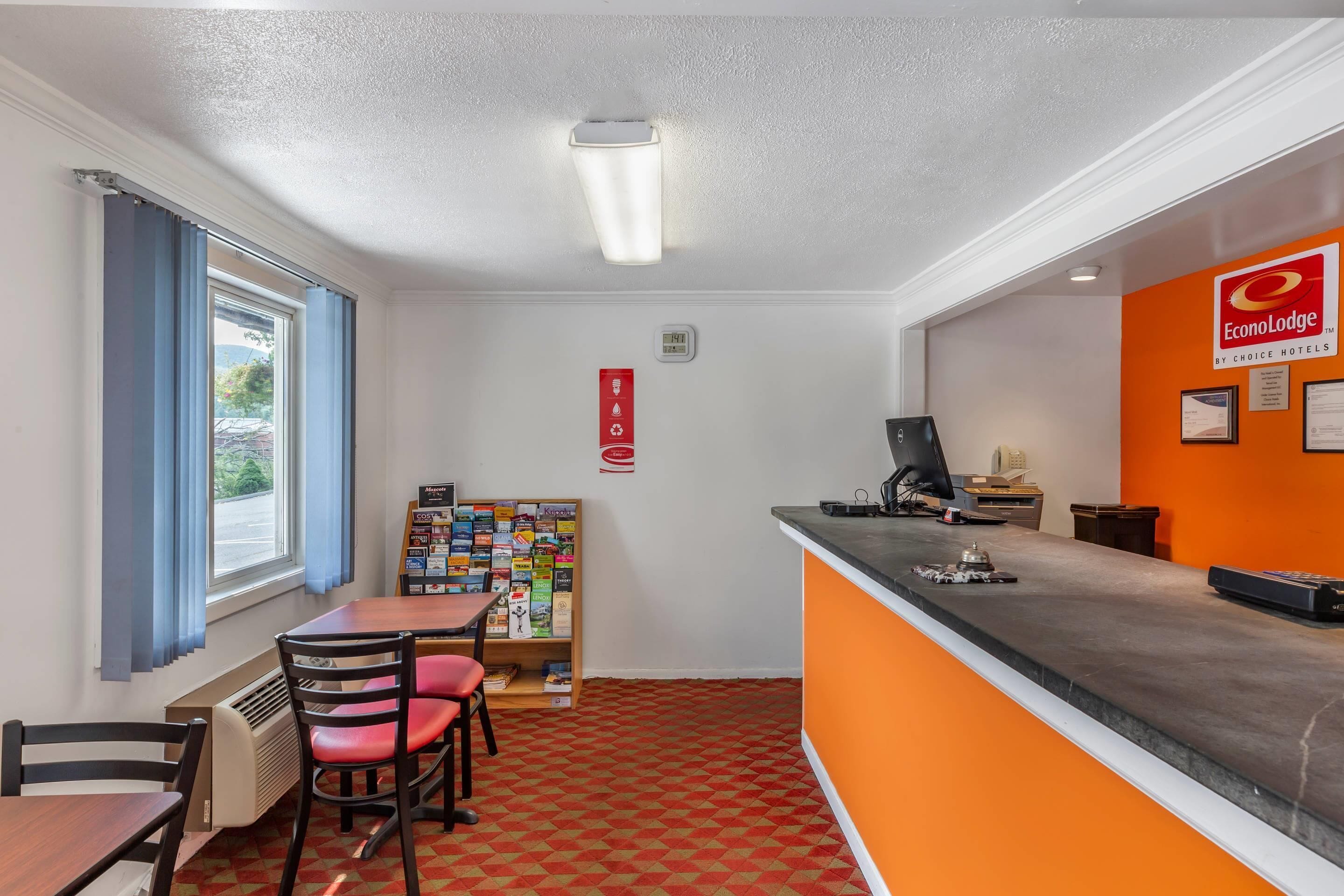 A hotel reception area with a desk, chairs, a bulletin board, and a window with curtains.