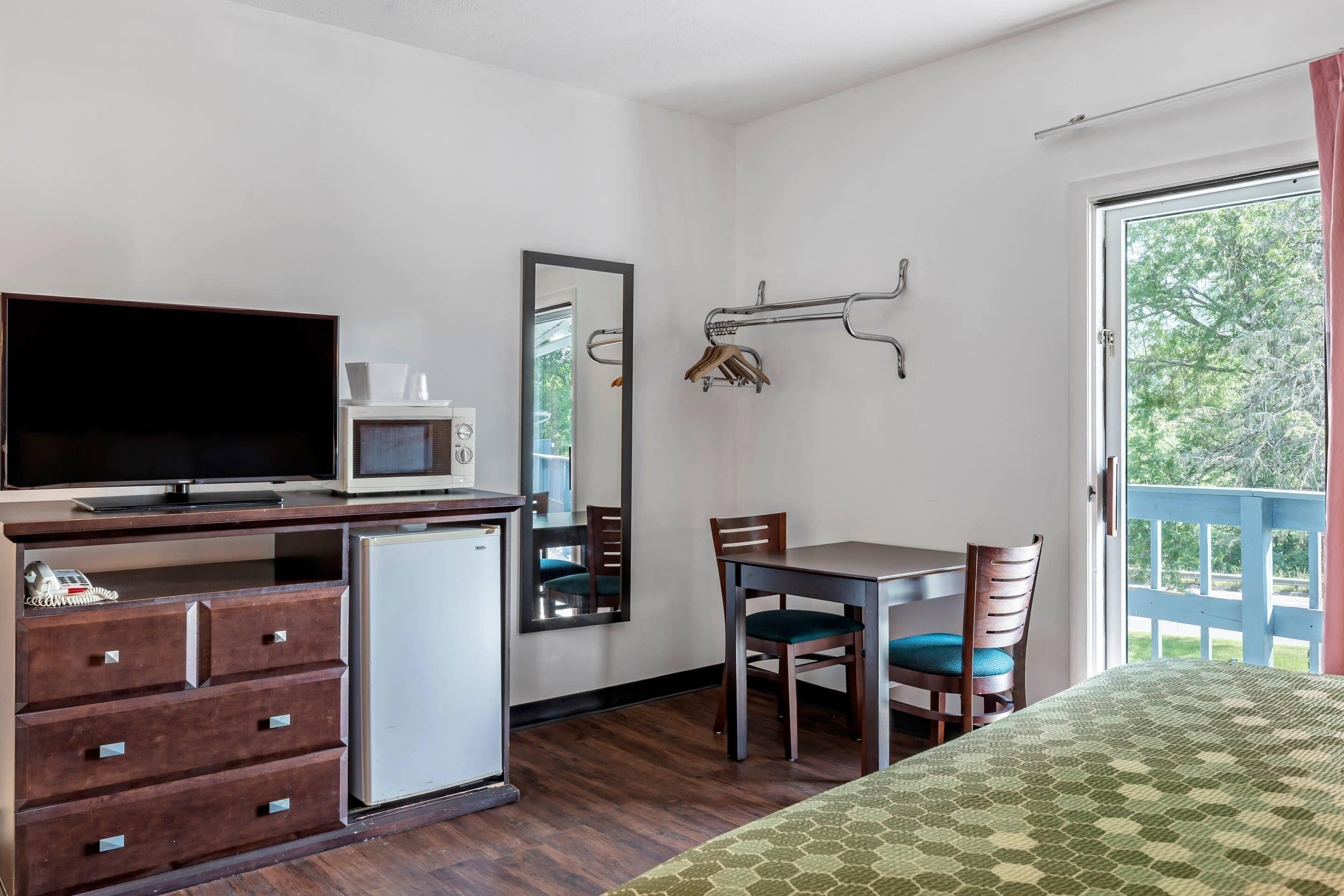 A compact room with a bed, a desk with a television, a small refrigerator, a mirror, and a window with a view of trees.