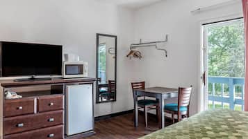 A compact room with a bed, a desk with a television, a small refrigerator, a mirror, and a window with a view of trees.