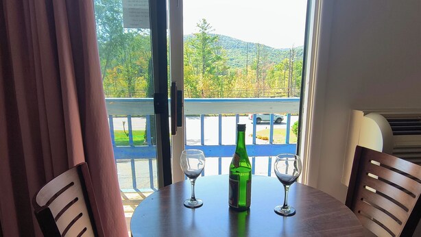 A round wooden table with two wine glasses and a green bottle, set against a window with a view of a blue railing and trees. - Econo Lodge Lee - Great Barrington (Lee)