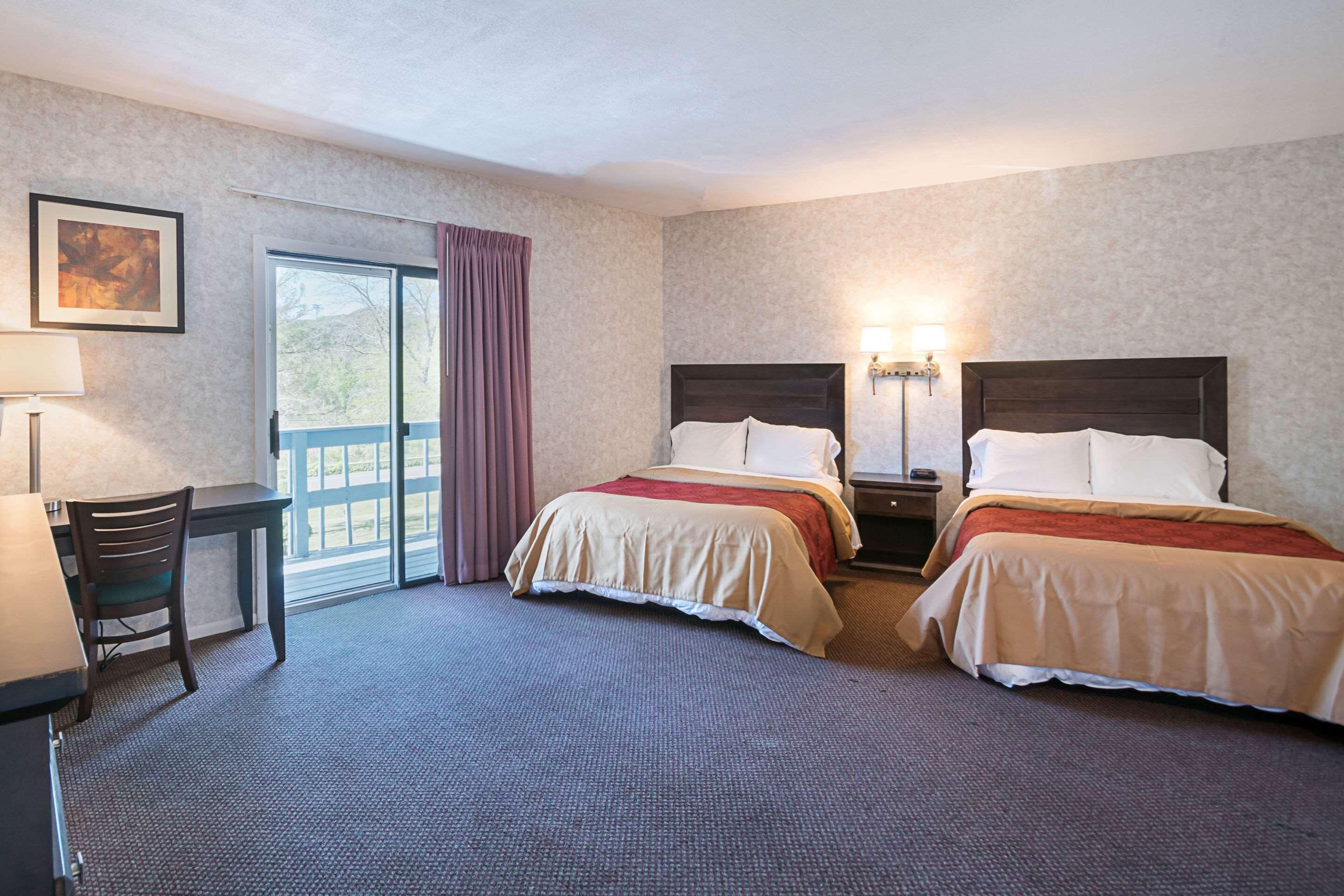 A hotel room with two beds, a desk, a chair, a window with curtains, and a wall-mounted lamp.