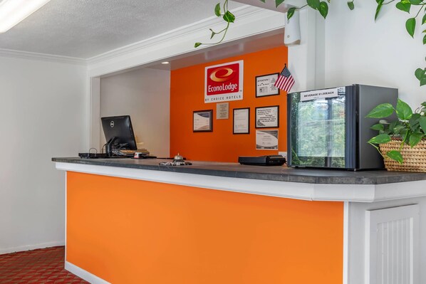 A hotel reception desk with an orange counter, a computer, a beverage cooler, and framed awards on the wall. - Econo Lodge Lee - Great Barrington (Lee)