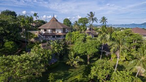 A large, multi-story house surrounded by lush greenery and palm trees, with a view of the ocean and distant mountains. - Mangosteen Ayurveda & Wellness Resort (Rawai)