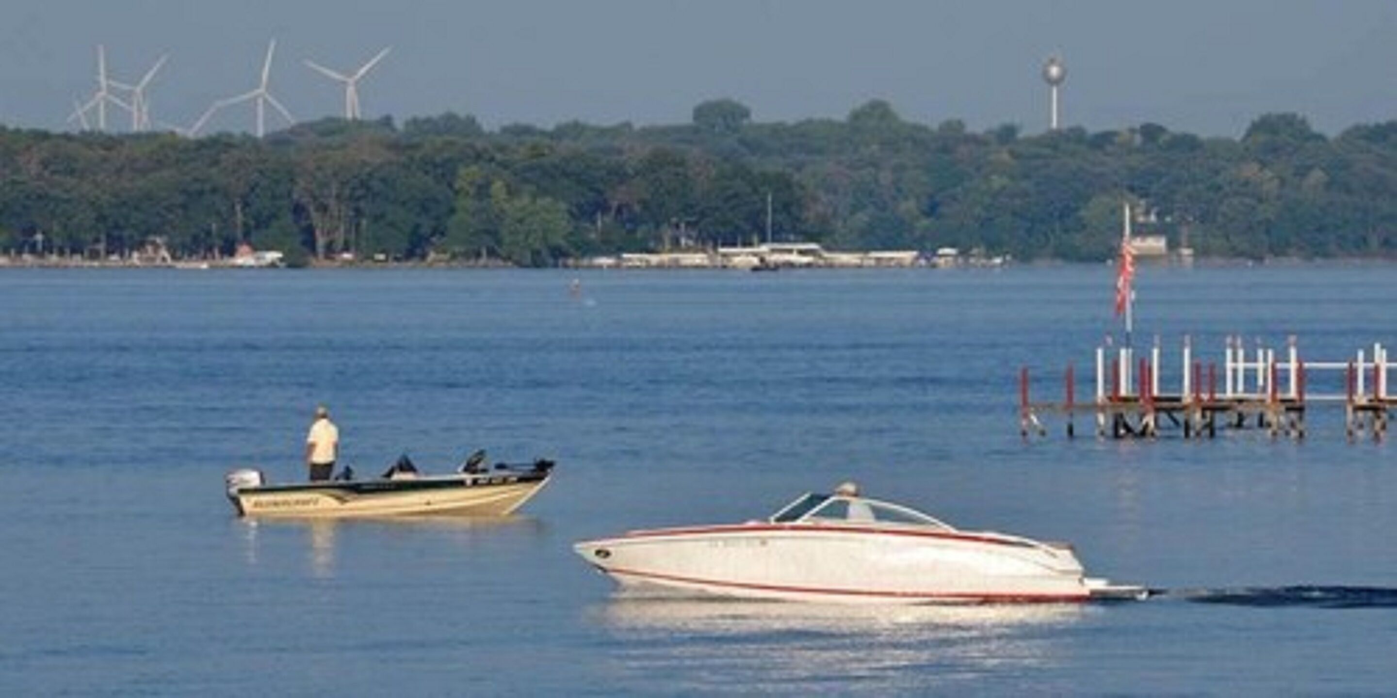 A boat on a calm lake with a dock and wind turbines in the background.