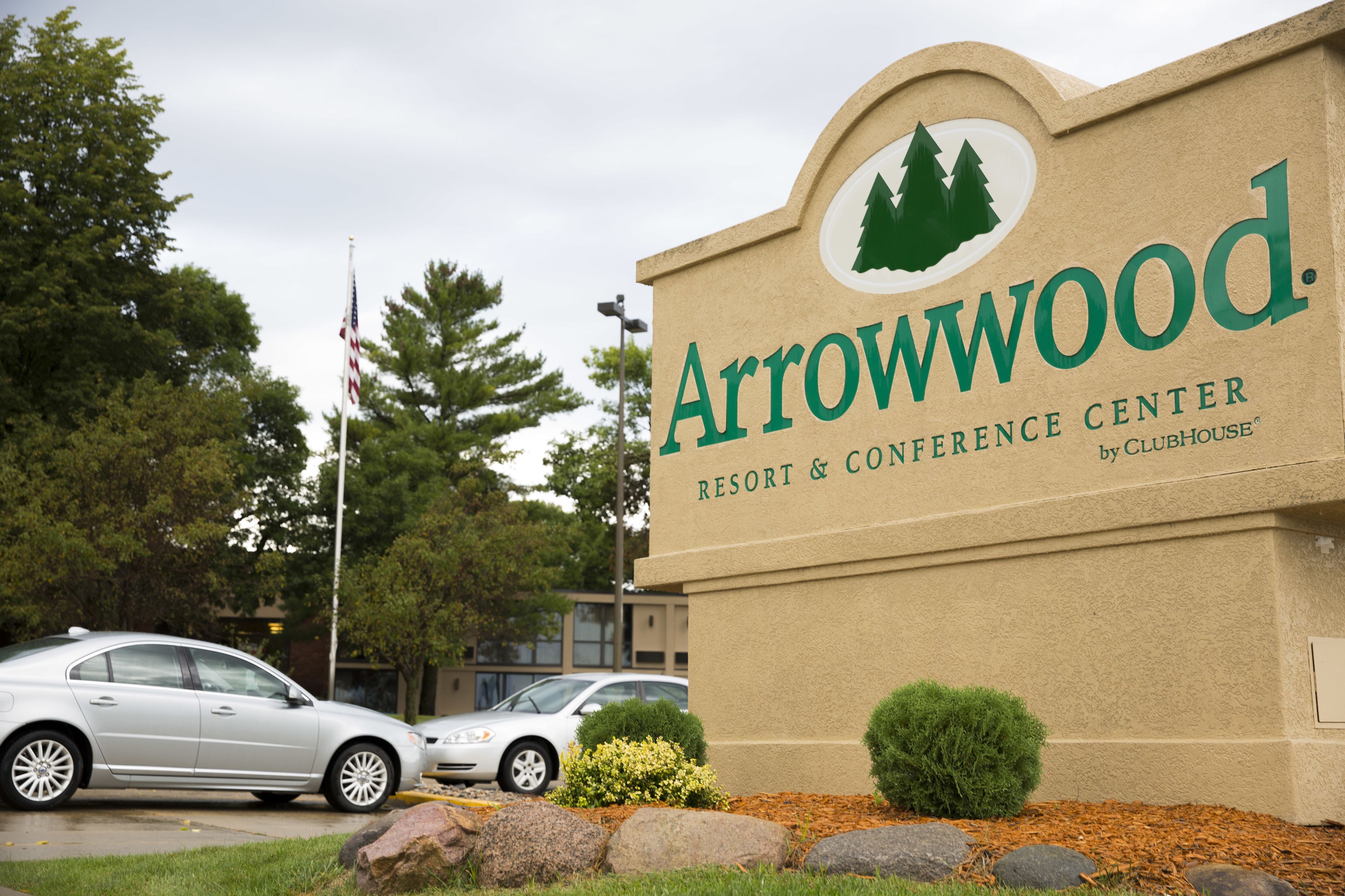 Señal del Arrowwood Resort & Conference Center por Clubhouse, con un logotipo que muestra árboles, un área de estacionamiento con coches y una bandera.