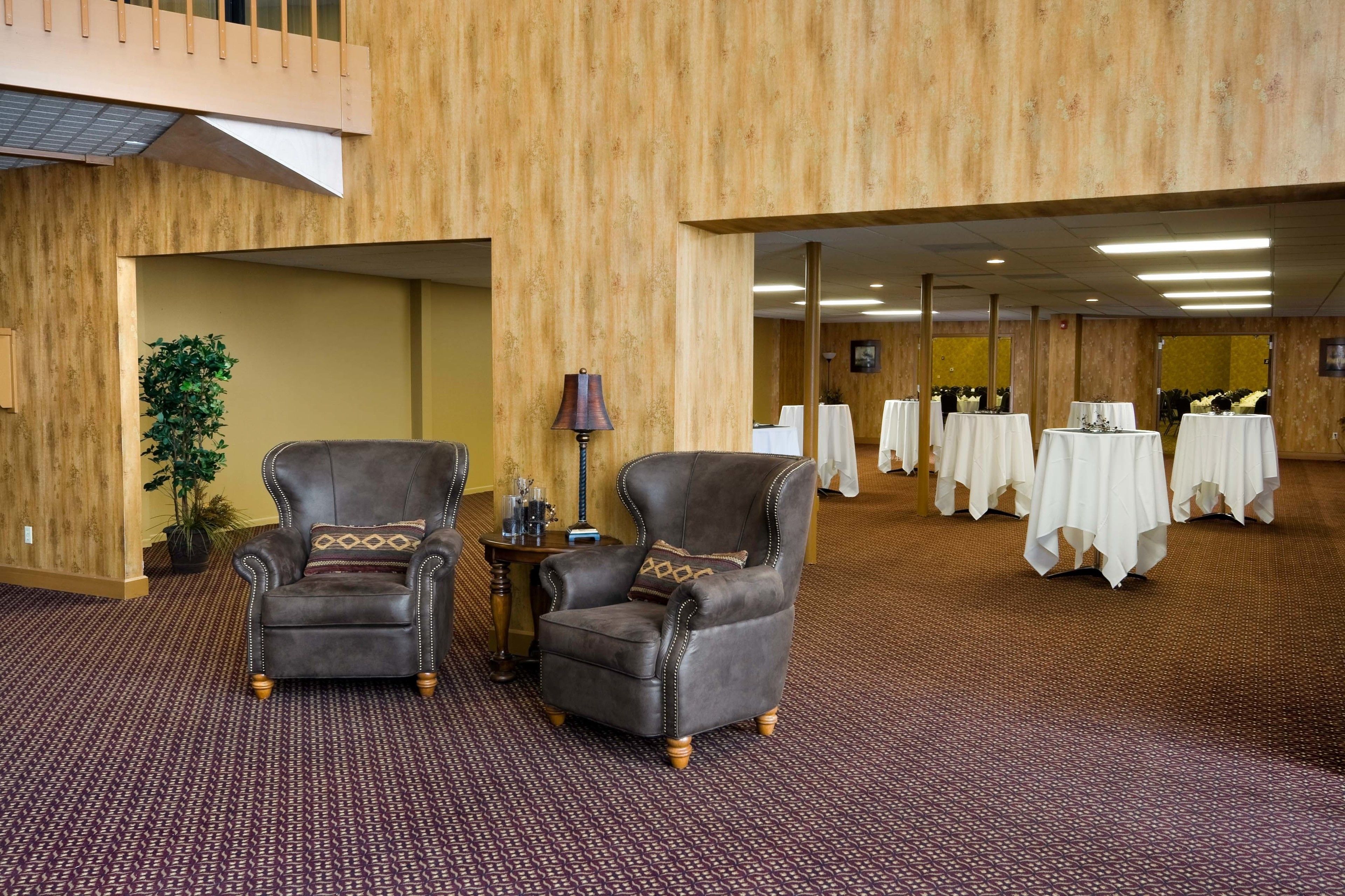 A hotel lobby with two leather armchairs, a small table with a lamp, and a large carpeted area with tables set up for dining.