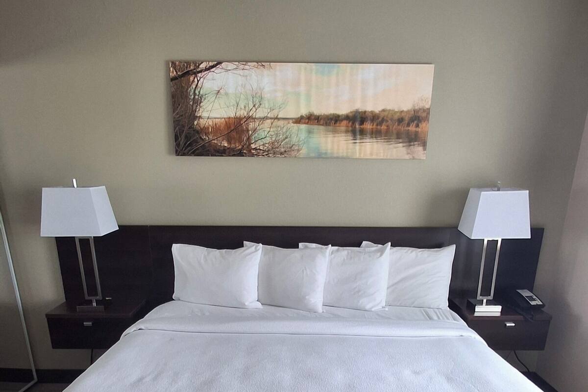 A large bed with white linens, two bedside tables with lamps, a painting on the wall, and a dark floor.