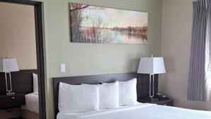 A hotel room with a large bed, two bedside tables with lamps, a painting on the wall, and a window with curtains. - Island Suites (Lake Havasu City)