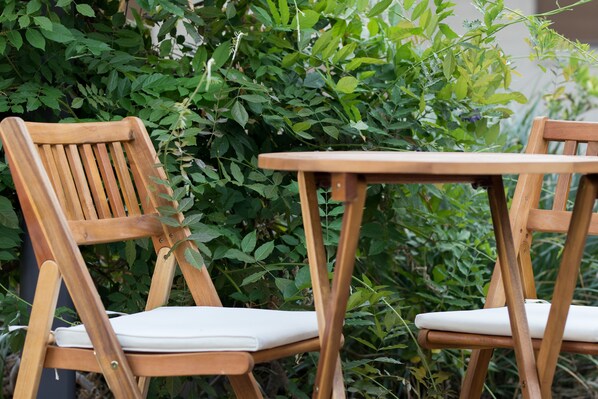 Deux chaises en bois avec des coussins blancs et une table en bois, disposées devant un décor de feuillage verdoyant et luxuriant.