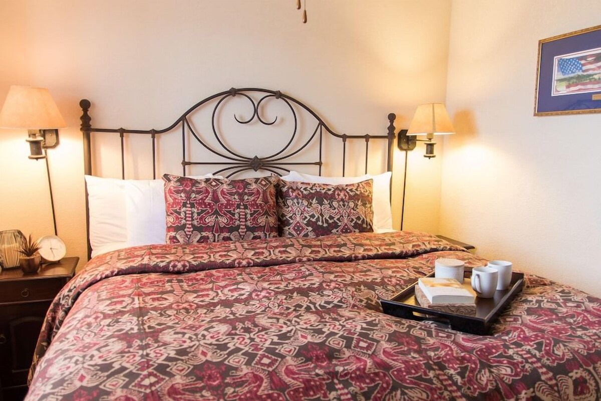 A neatly made bed with patterned bedding, two bedside lamps, a tray with a book and cups, and a framed picture on the wall.