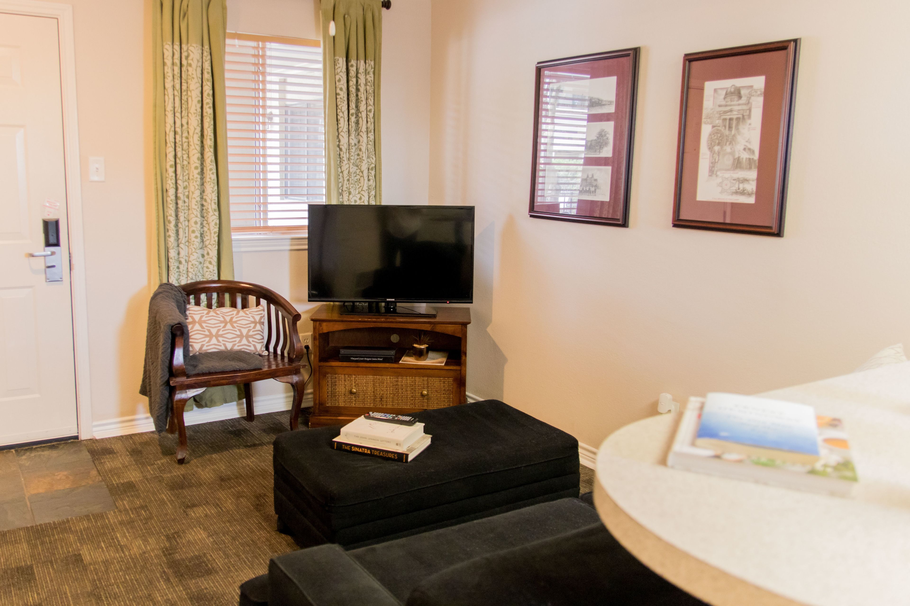 Une chambre d’hôtel équipée d’une télévision, d’une chaise, d’un canapé, d’une petite table avec des livres et de deux tableaux encadrés accrochés au mur.