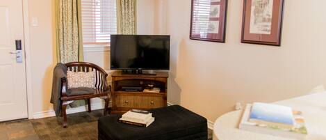 A hotel room with a television, a chair, a sofa, a small table with books, and two framed pictures on the wall.