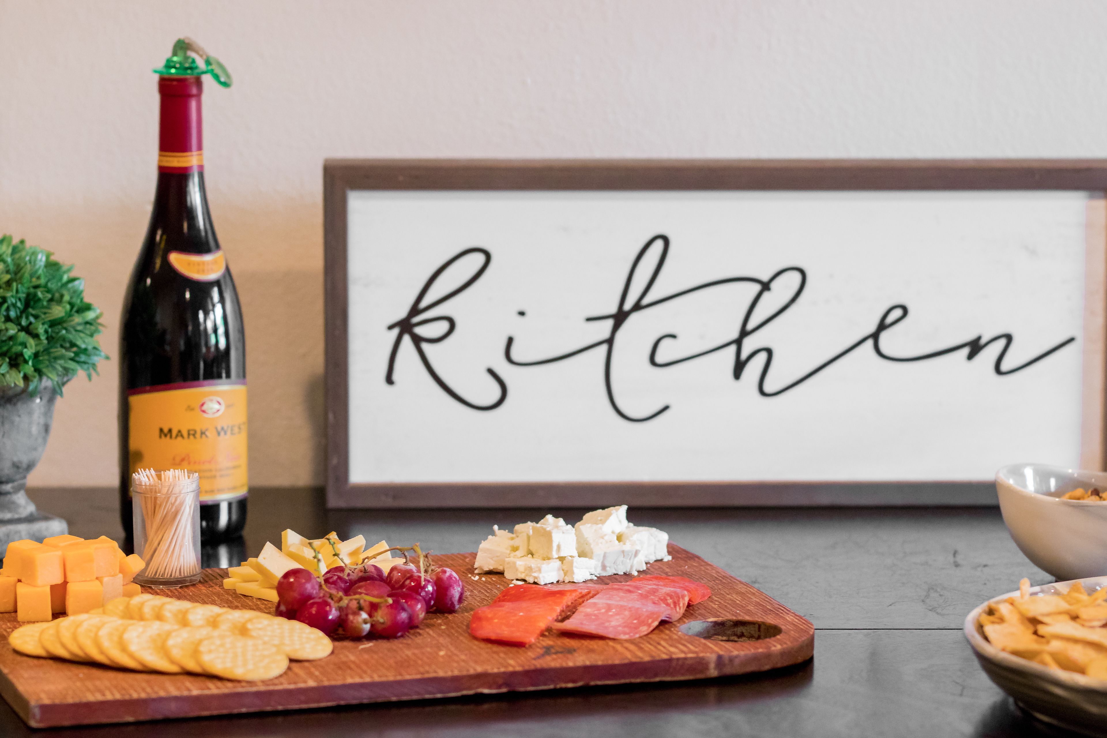 Ein Holzbrett mit Käse, Crackern, Trauben und Aufschnitt, eine Weinflasche und ein gerahmtes Schild mit dem Wort 'kitchen'.