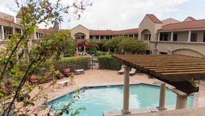Zona de piscina con sillones de descanso y una pérgola, rodeada por un patio con un edificio al fondo.