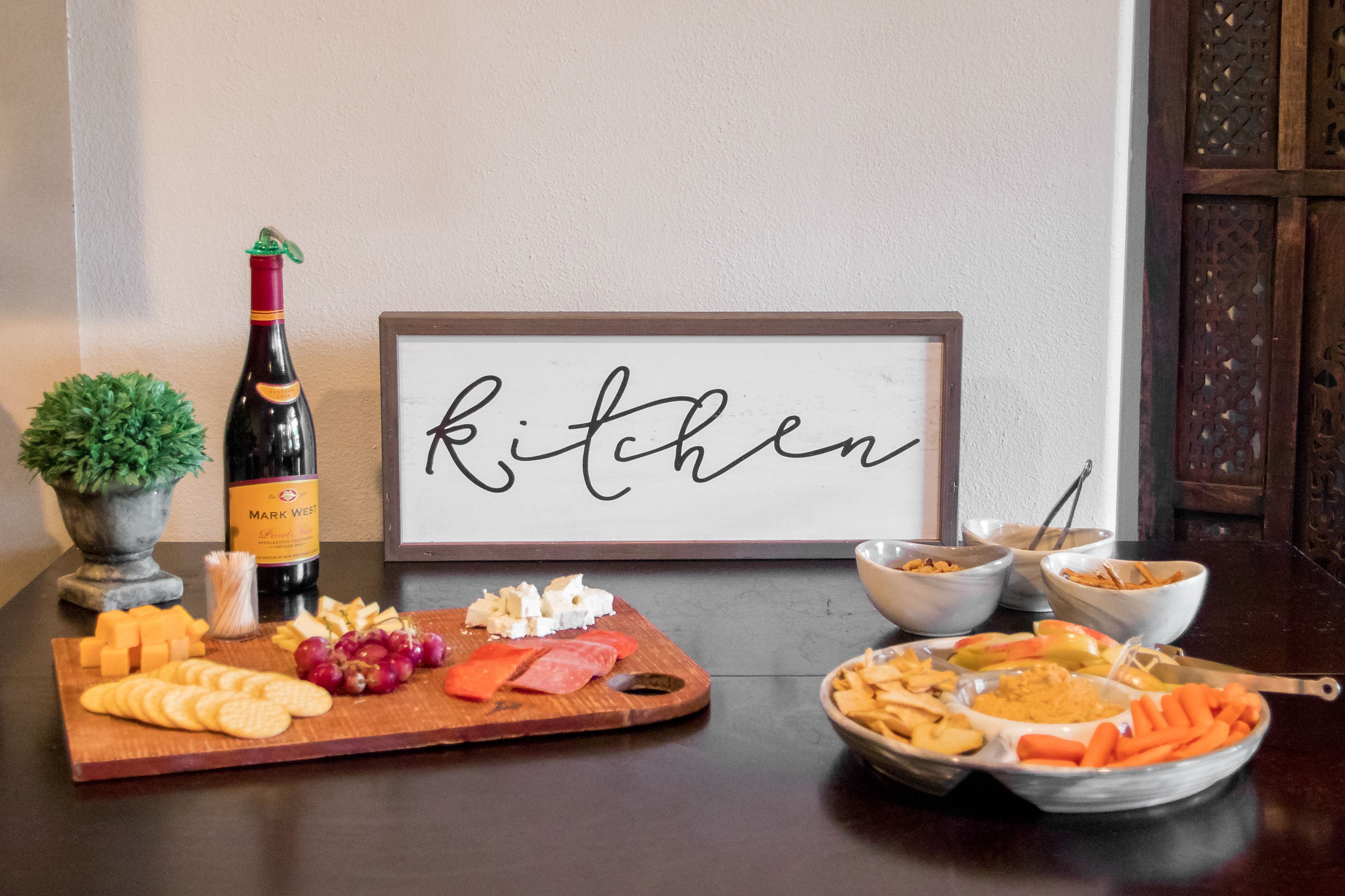 A table with a variety of cheeses, crackers, and fruits, a bottle of wine, and a sign that reads "kitchen".
