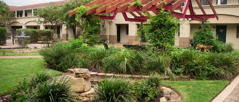 Un patio con una pérgola, abundante vegetación y un edificio con techo de tejas rojas.