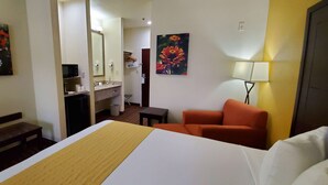 Ein Hotelzimmer mit einem Bett, einer Bank, einem Schreibtisch, einem Sessel, einem Spiegel, einem Waschbecken und zwei Bildern.