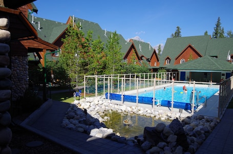 A pool area with a small pond, surrounded by stone walls and wooden buildings with green roofs.