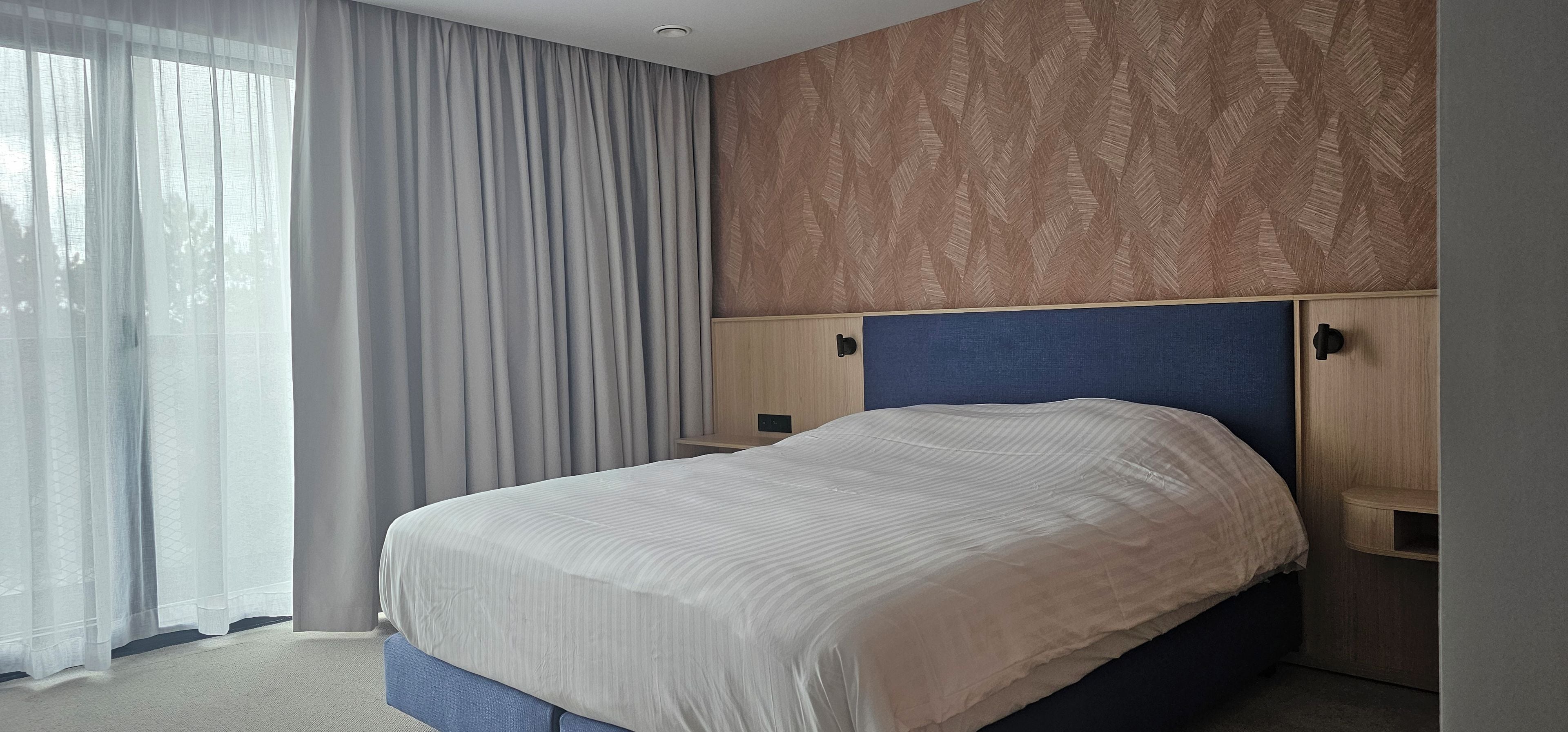 Habitación de hotel con cama, mesita de noche, lámpara de pared y ventana con cortinas translúcidas.