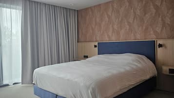 A hotel room with a bed, a nightstand, a wall-mounted lamp, and a window with sheer curtains.