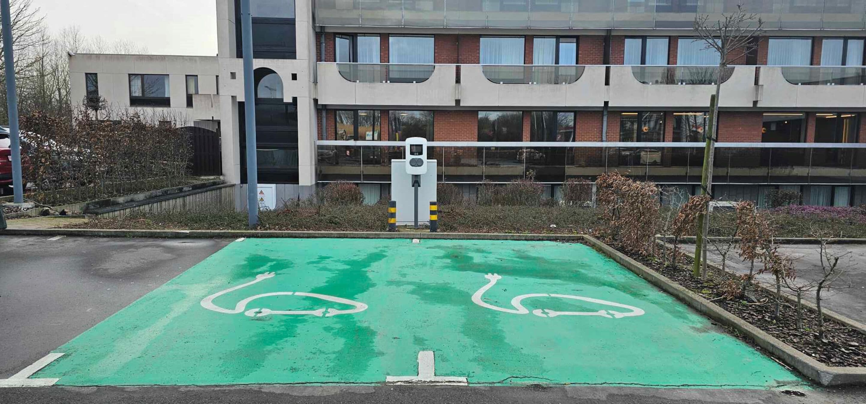 Un’area di parcheggio con spazi verdi e posti auto riservati ai disabili, un edificio con ampie finestre e una colonnina di ricarica elettrica ben visibile.
