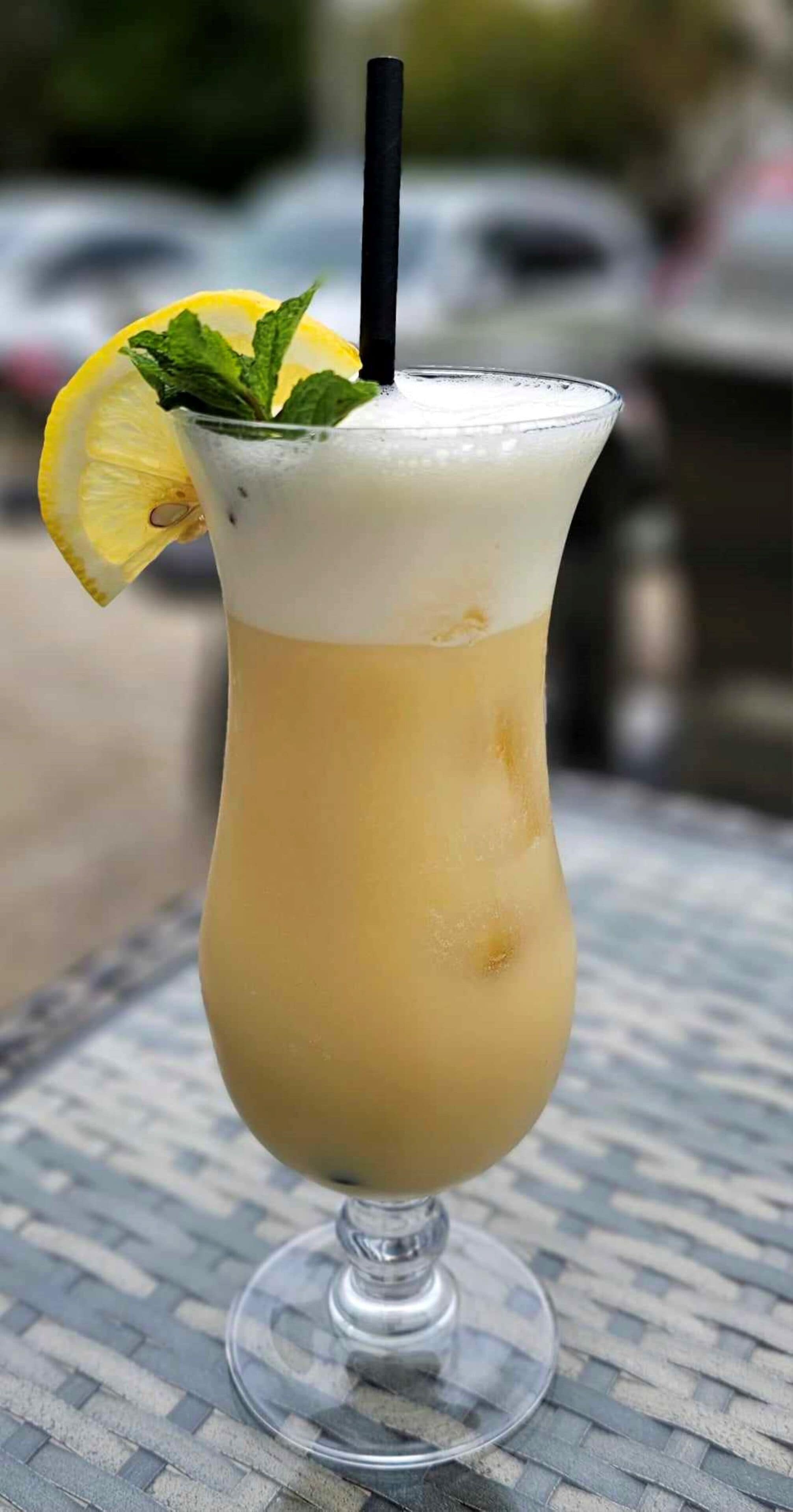 Un bicchiere alto con una cannuccia, contenente una bevanda chiara, guarnito con una fetta di limone e foglie di menta, su un tavolino intrecciato.