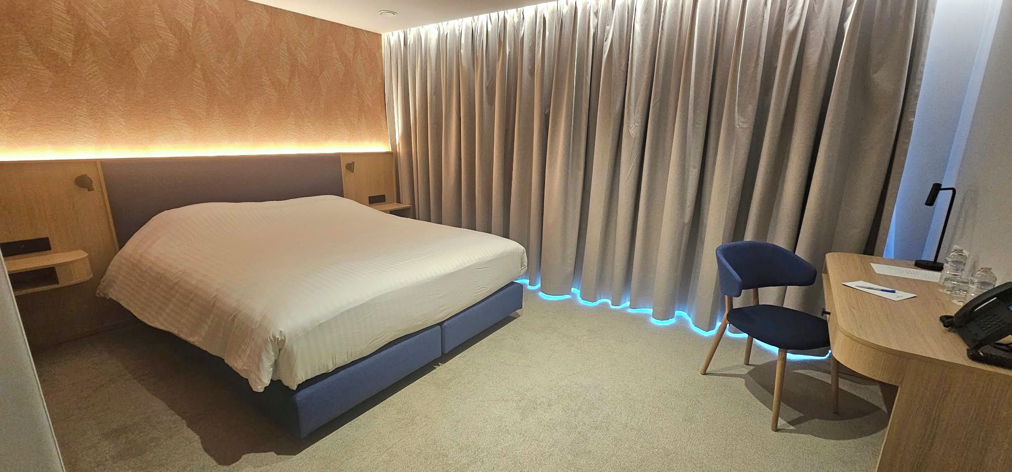 A hotel room with a bed, a desk with a chair, and a curtain with blue LED lighting.