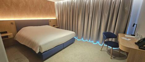 Habitación de hotel con cama, escritorio con silla y cortina con iluminación LED azul.