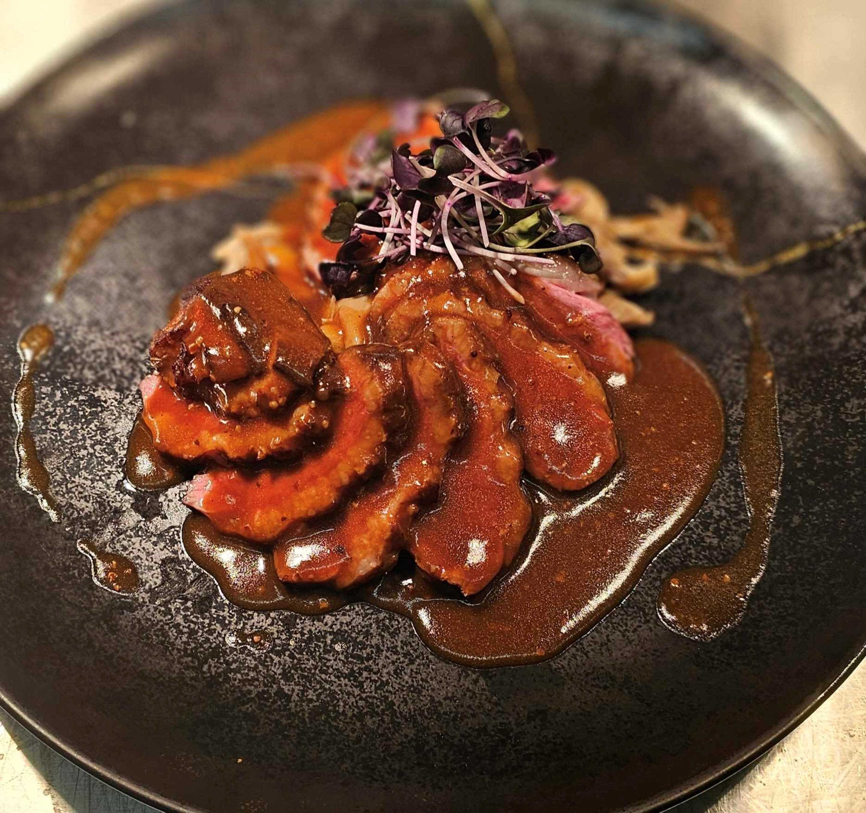 Une assiette de nourriture comprenant de la viande coupée en tranches et une sauce, le tout agrémenté de feuilles violettes.