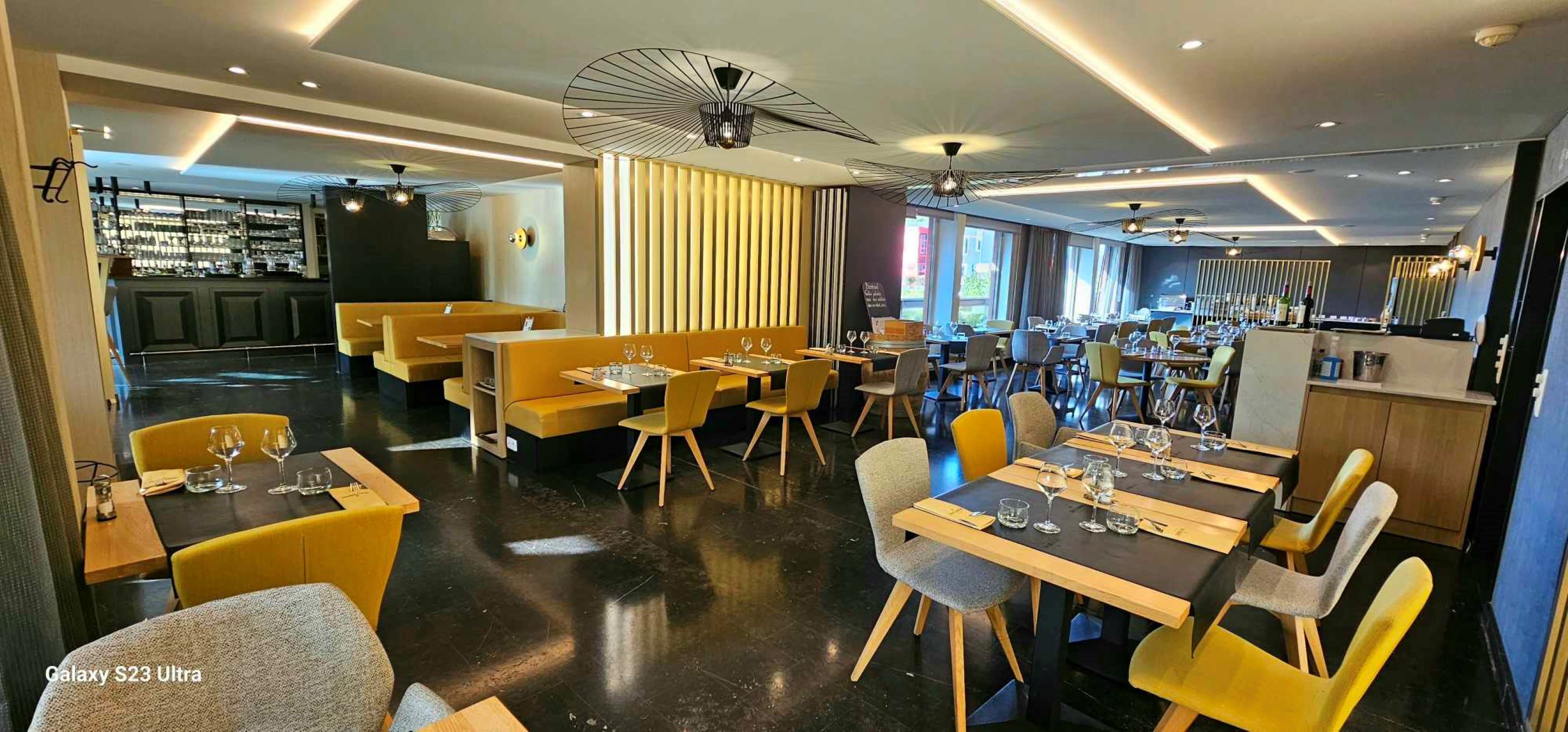 Ein modernes Restaurant mit gelben Sitzbänken und Tischen, einer Bar und Blick auf die Stadt.