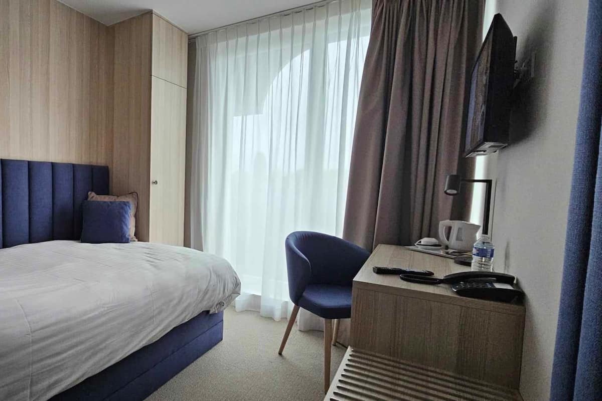 A hotel room with a large bed, a desk with a chair, a flat-screen TV, and a window with curtains.