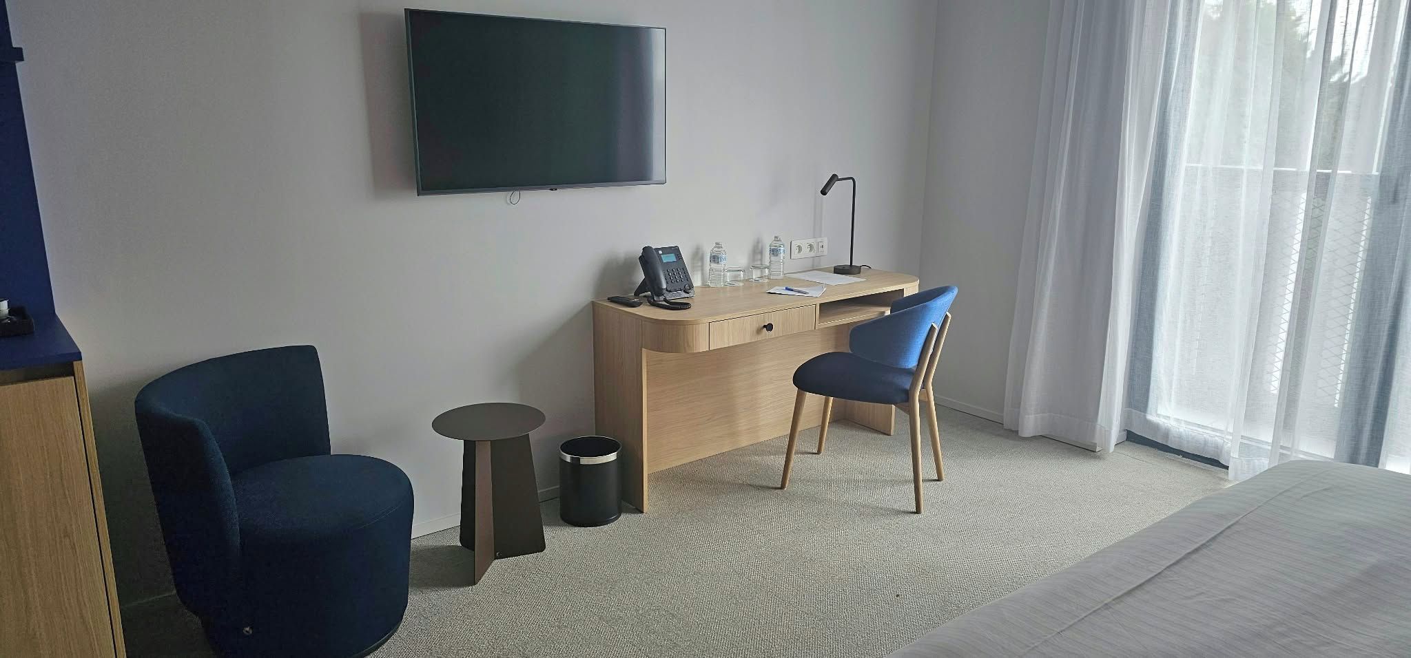 A hotel room with a bed, a desk with a chair, a television, and a window with curtains.
