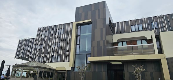 Un edificio moderno con paneles verticales y horizontales, ventanas amplias y un área de estar al aire libre cubierta.