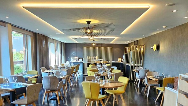 A modern restaurant with a central dining area, high ceiling with a unique light fixture, and a view of the outdoors. - Hotel Les 3 Clés (Gembloux)