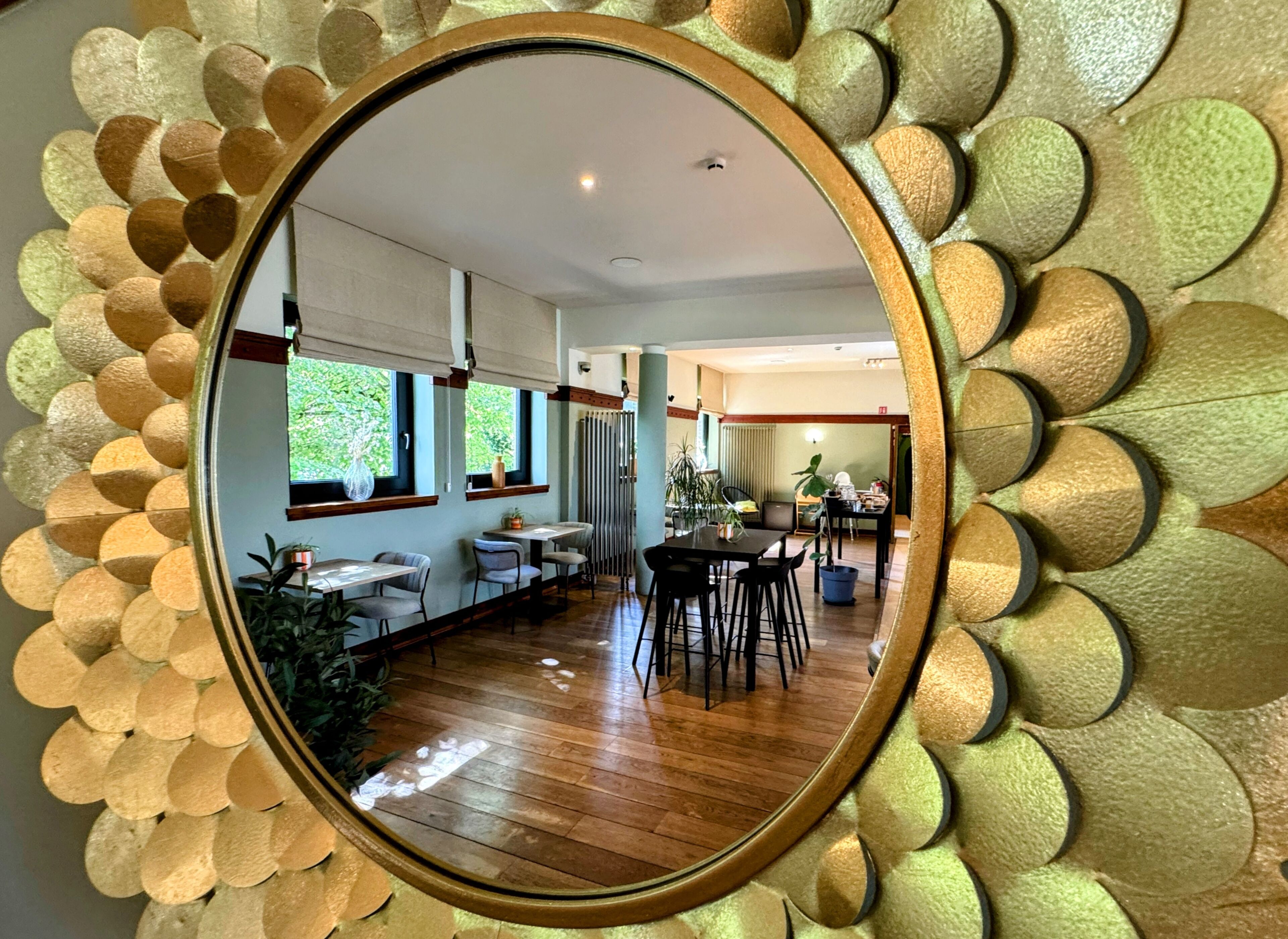 A room with a wooden floor, a dining area with tables and chairs, and a view of a kitchen area through a mirror.