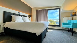 Habitación de hotel con una cama grande, una ventana con cortinas, vistas a la ciudad, una silla azul y un radiador.