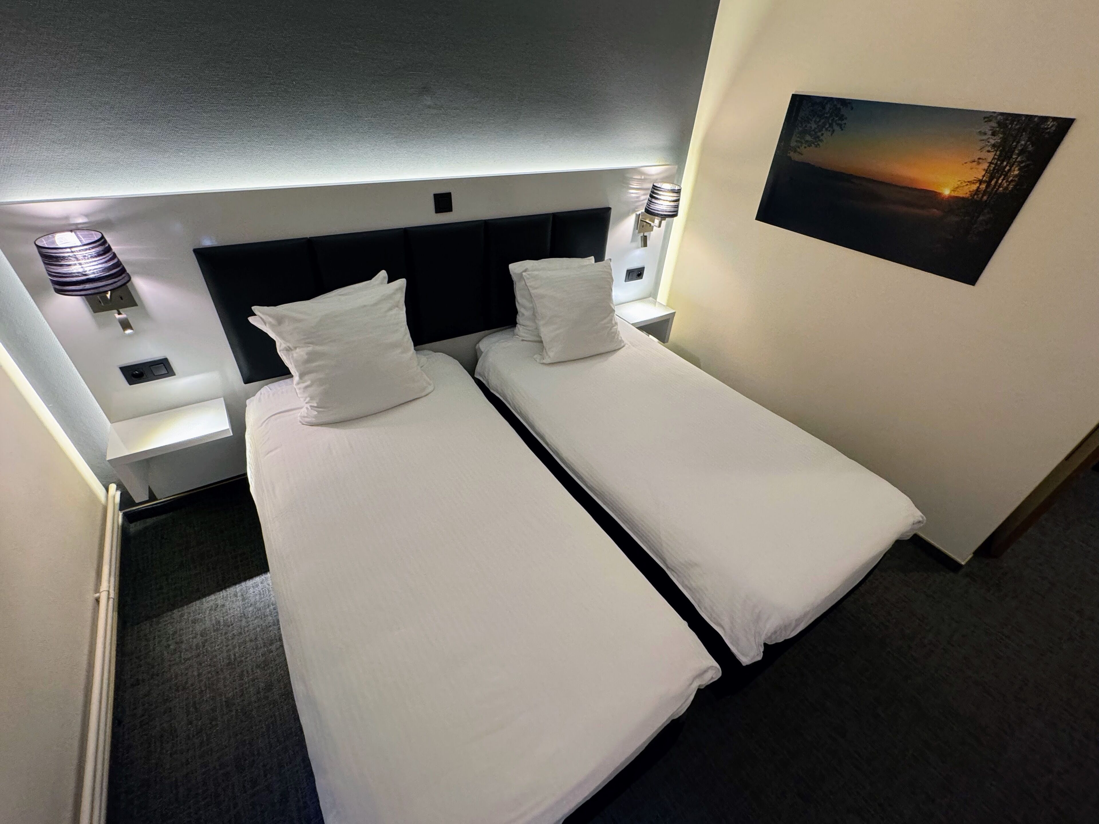 Una habitación de hotel con dos camas individuales, cabeceras, lámparas de noche, una mesita y un cuadro en la pared.