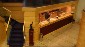 Un mostrador de recepción con un hombre detrás, una escalera a la izquierda y un basurero a la derecha.