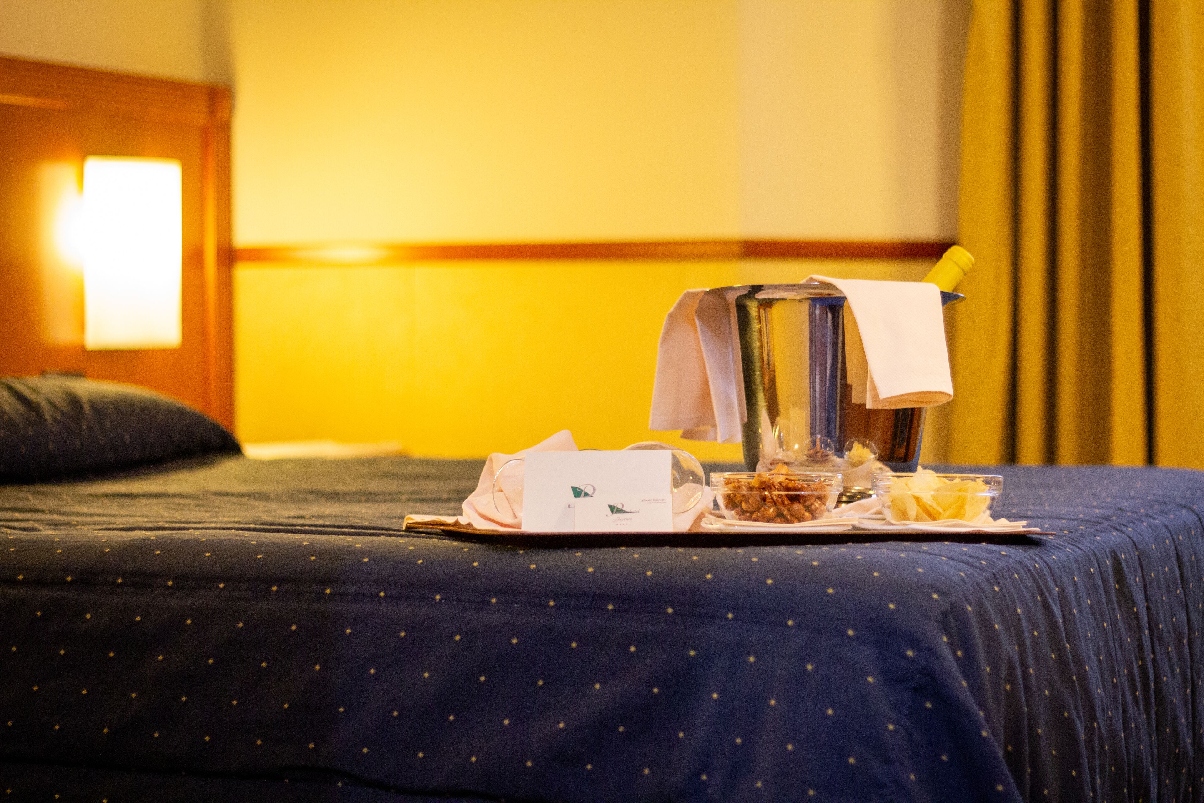 A neatly made bed with a dark blue bedspread and a tray with breakfast items, including cereal and a cup, on a hotel room.
