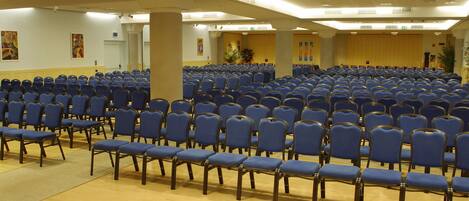 A large hall with rows of blue chairs and wooden floors, likely a conference or meeting room.