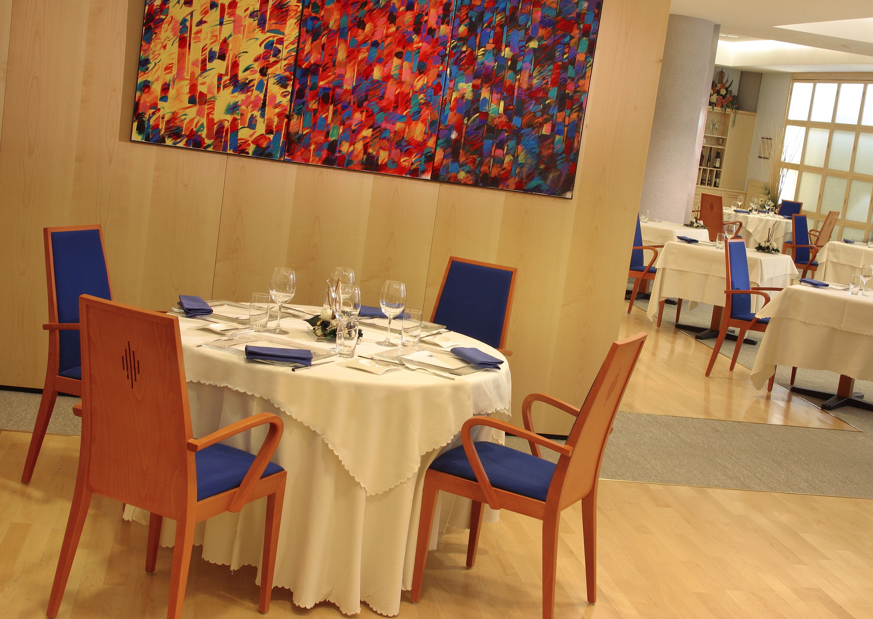 Un comedor con mesas redondas, sillas azules y un cuadro abstracto grande en la pared.