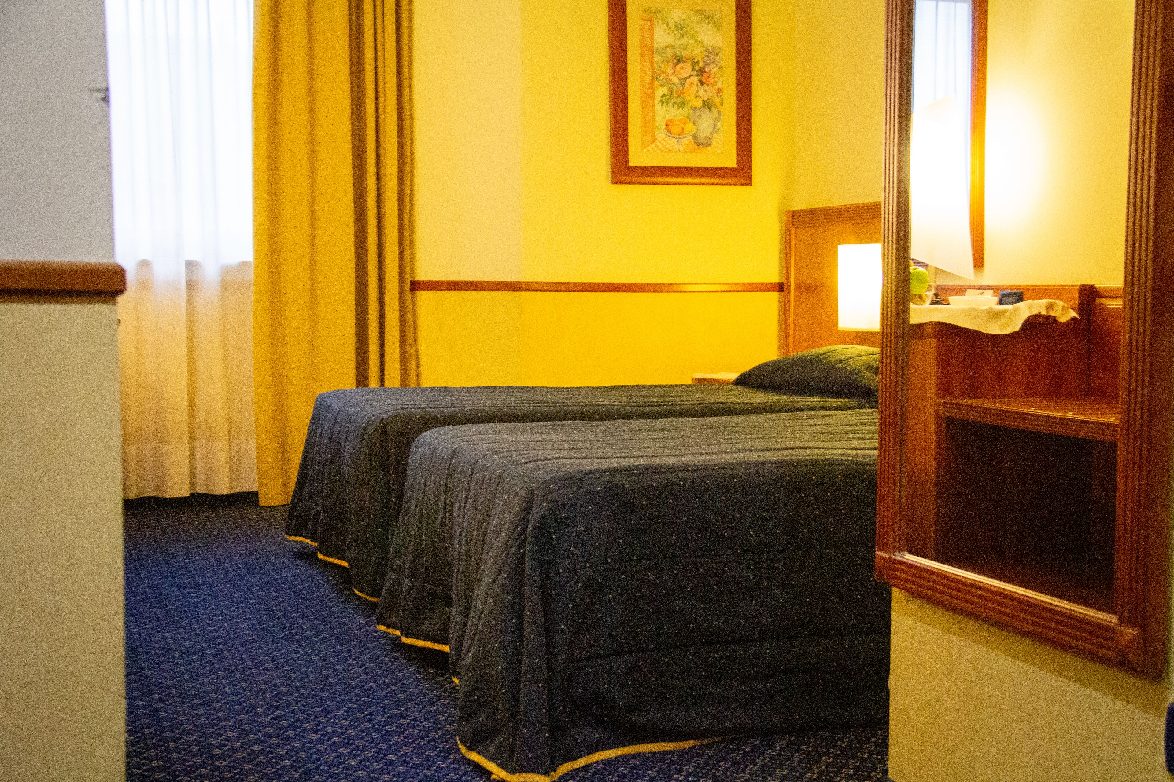 A hotel room with two beds, a large mirror, a painting on the wall, and a window with curtains.