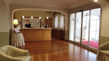 Ein Hotel-Empfangsbereich mit einem Holzbüro, einer Rezeptionistin und einem großen Fenster, wobei ein roter Teppich nach draußen führt.