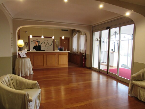 A hotel reception area with a wooden reception desk, a receptionist, and a large window with a red carpet leading outside.