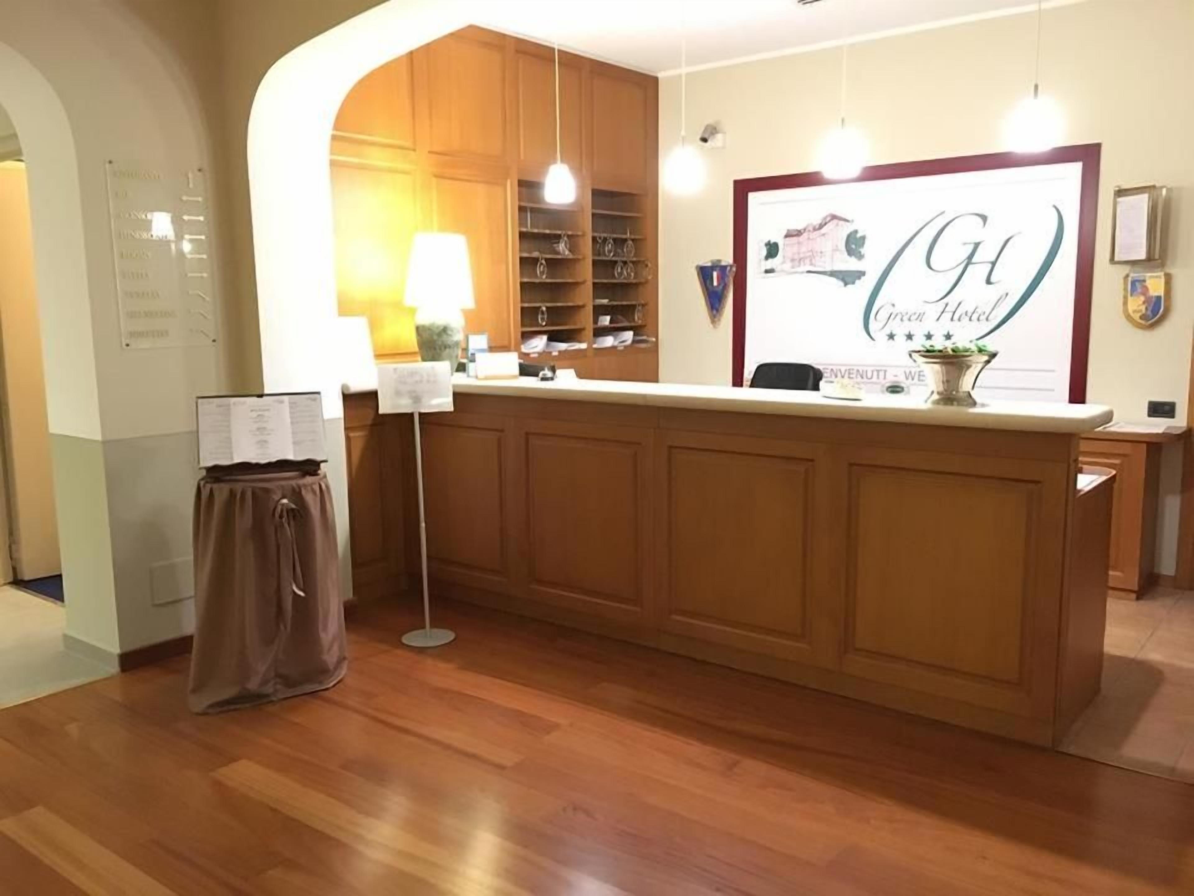 A hotel reception area with a wooden reception desk, a framed sign above it, and a wall-mounted information board.
