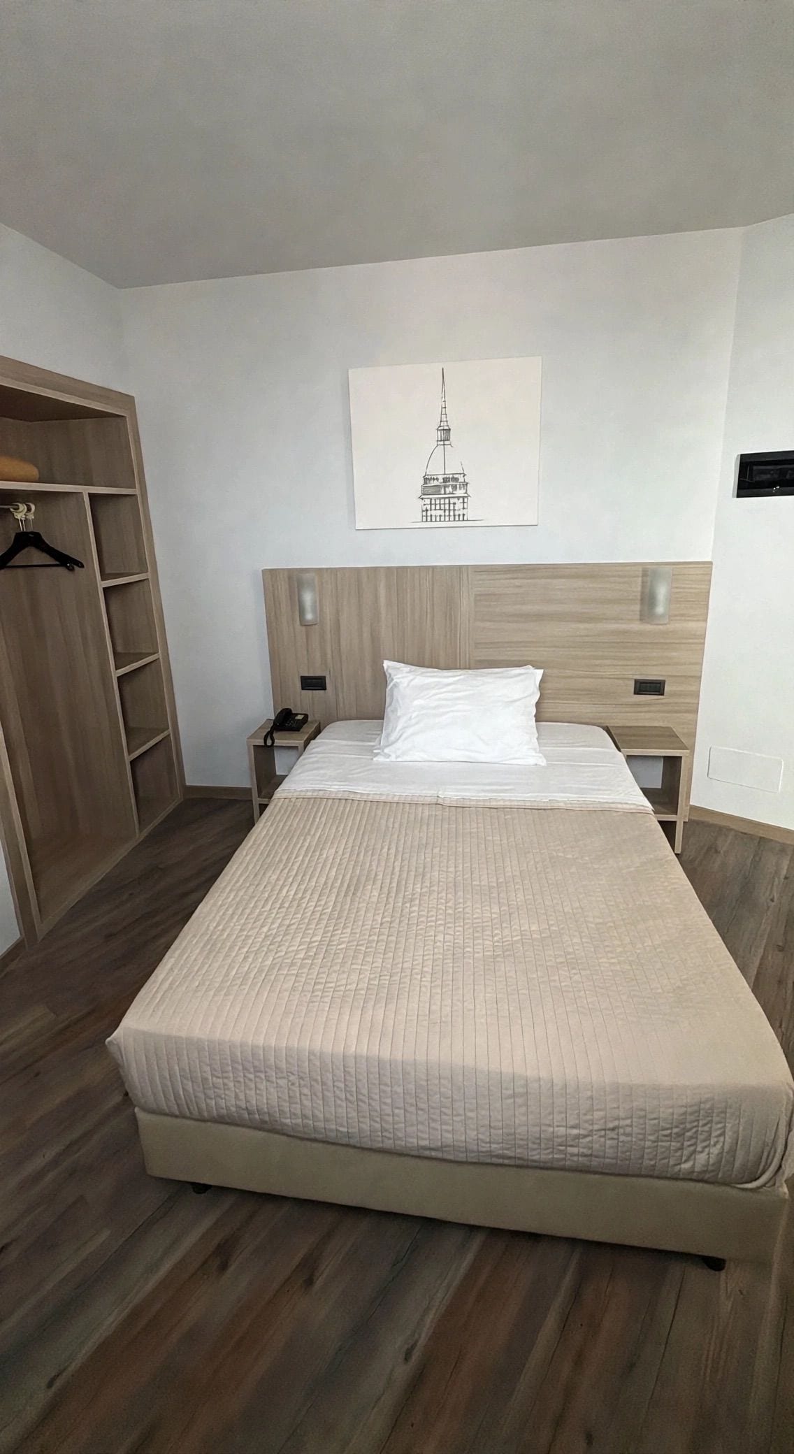 A hotel room with a large bed, a wooden wardrobe, a bedside table with a phone, and a wall-mounted light.