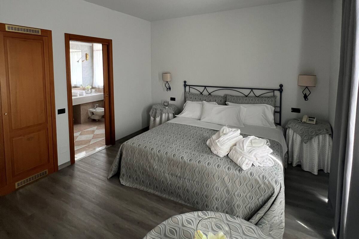 A hotel room with a large bed, two bedside tables, a round table with a vase, and a mirror in the corner.