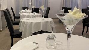 Een eetruimte met ronde tafels, elk gedekt met een witte tafelkleed, wijnglazen en een gevouwen servet.
