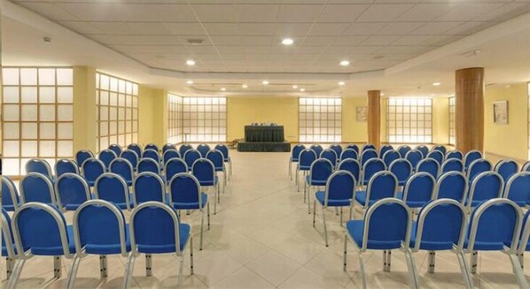 A conference room with rows of blue chairs, a stage, and large windows.