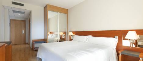 A hotel room with a large bed, wooden headboard, bedside tables with lamps, a mirror, and a wooden bench.